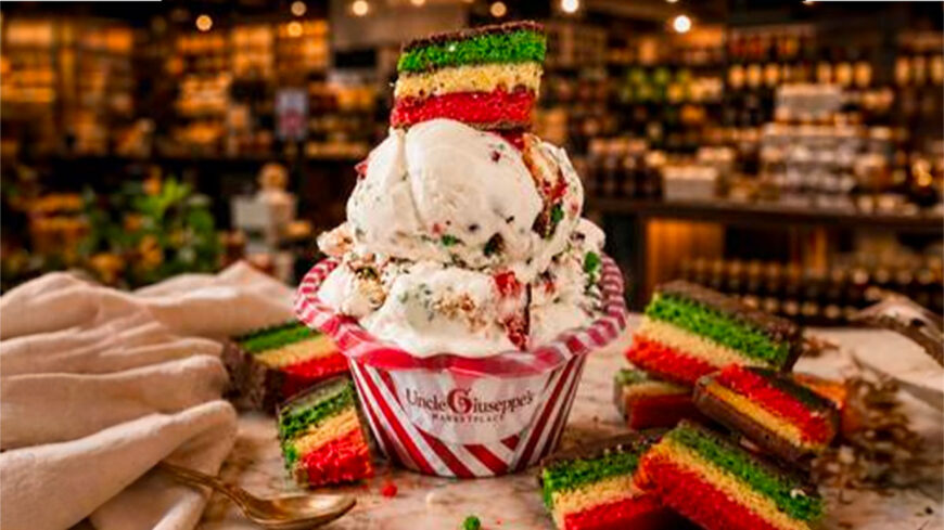 Two scoops of vanilla ice cream with colorful cake pieces, topped with a slice of Italian rainbow cake, served in a striped Uncle Giuseppe's Marketplace cup. Additional rainbow cake slices surround the dessert on a table, with a grocery market blurred in the background.
