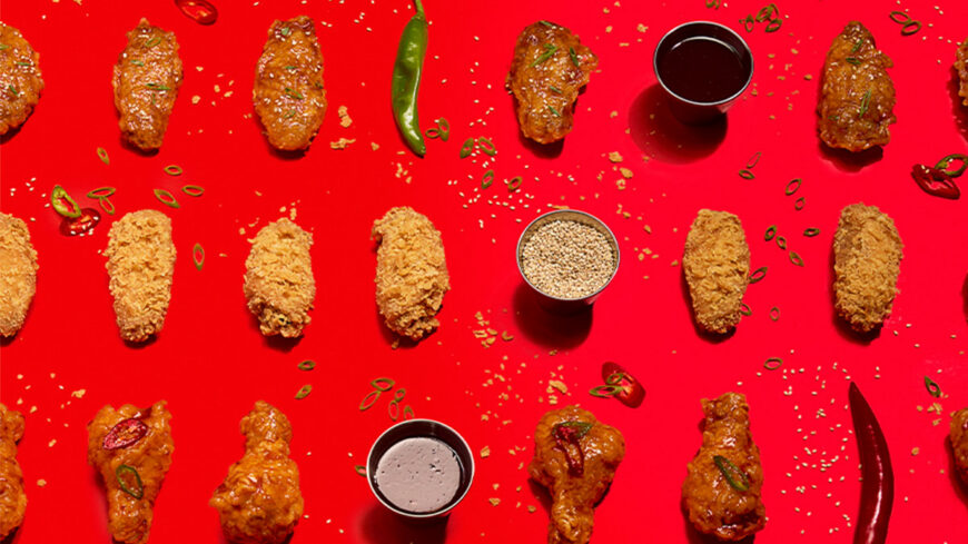 An assortment of crispy fried and sauced chicken wings arranged in a neat grid on a bright red background, with dipping sauces, chili peppers, sesame seeds, and sliced green onions scattered throughout.
