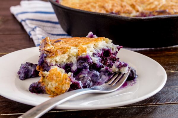 Close up of slice of fresh baked blueberry cobbler sitting on white plate with fork