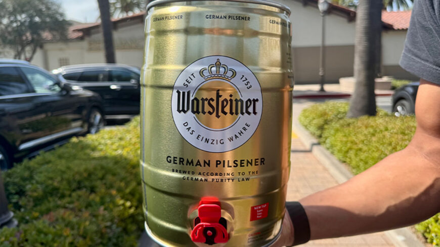 A person holds a Warsteiner German Pilsener mini keg with a red tap outdoors.