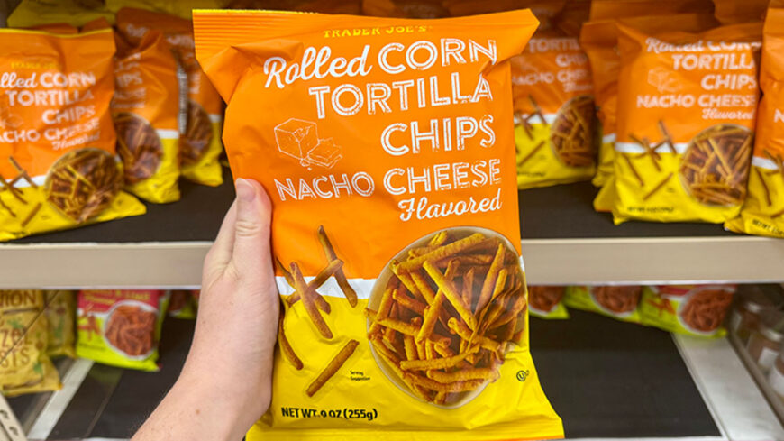 A hand holds a bag of Trader Joe’s Rolled Corn Tortilla Chips flavored with Nacho Cheese on a grocery store shelf.
