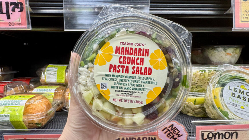 A hand holds a container of Trader Joe’s Mandarin Crunch Pasta Salad with mandarin oranges, apples, feta cheese, cranberries, and pumpkin seeds.