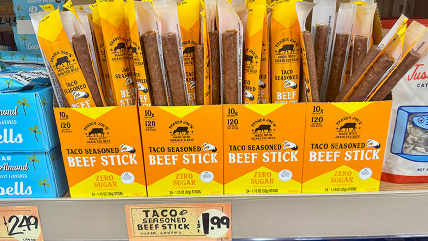 Trader Joe’s Taco Seasoned Beef Sticks in yellow-and-orange packages, arranged in a grocery store display with a $1.99 price sign.