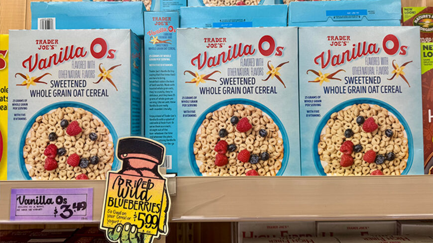 Boxes of Trader Joe’s Vanilla O’s sweetened whole grain oat cereal with fruit pictured on the front, displayed on a grocery store shelf with a $3.49 price sign.