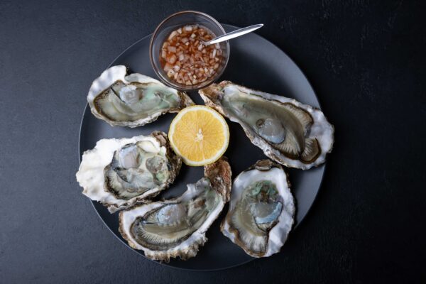 Khasan oysters, live, fresh, opened. Served with classic French Mignonette sauce and half a lemon. Blue plate, black background. Selective focus, close-up.