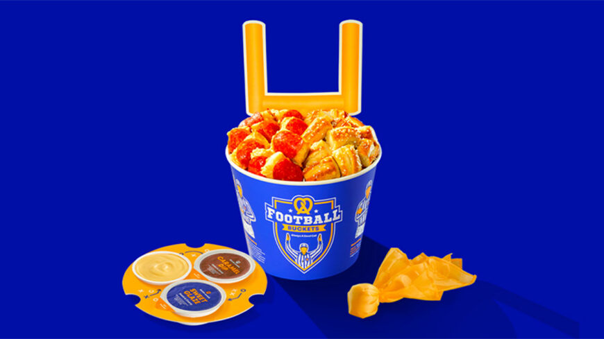 Auntie Anne’s Football Bucket filled with assorted soft pretzel bites and dipping sauces, set against a blue background with a large yellow football goalpost behind it.