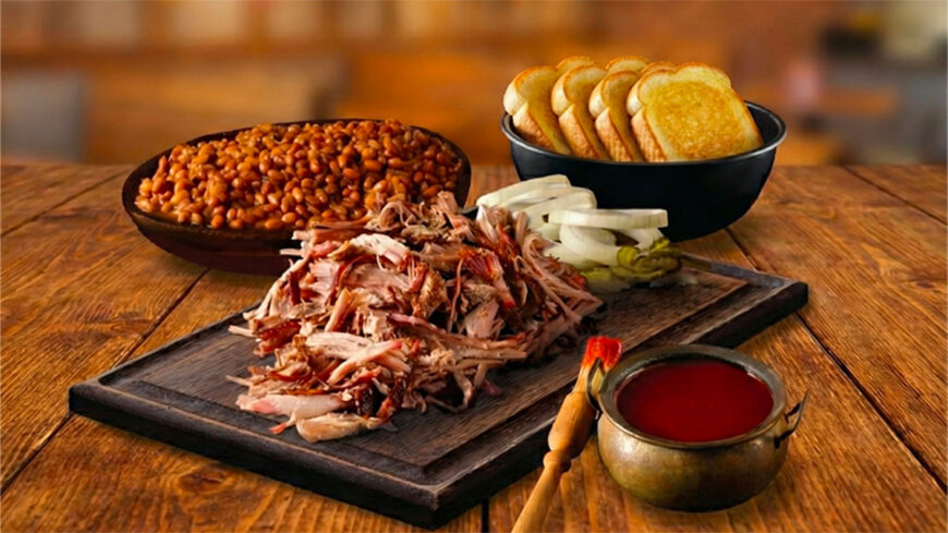 Barbecue spread featuring a pile of pulled pork, baked beans, sliced white bread, onions, pickles, and a bowl of red barbecue sauce on a wooden table.