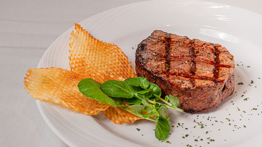 Grilled filet mignon with crosshatch grill marks, served with a crisp Parmesan cheese tuile and fresh greens on a white plate.