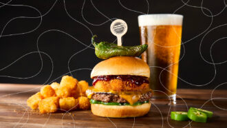 Cheeseburger topped with a crispy fried patty, barbecue sauce, and roasted green pepper beside tater tots and a beer, overlaid with white looping lines