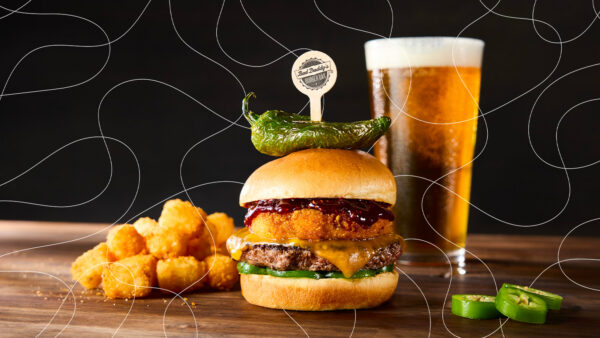 Cheeseburger topped with a crispy fried patty, barbecue sauce, and roasted green pepper beside tater tots and a beer, overlaid with white looping lines