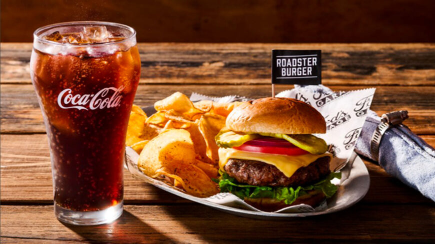 Cheeseburger with lettuce, tomato and pickles served with crispy chips and a glass of Coca-Cola on a wooden table.
