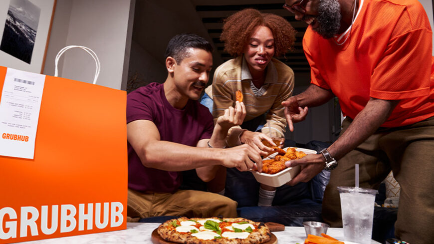 Three people sharing takeout food at home, with a Grubhub delivery bag in the foreground and pizza and wings on the table.