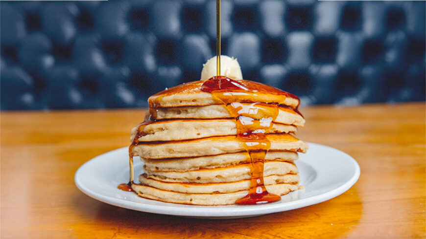 Stack of fluffy pancakes topped with butter and drizzled with maple syrup on a white plate.