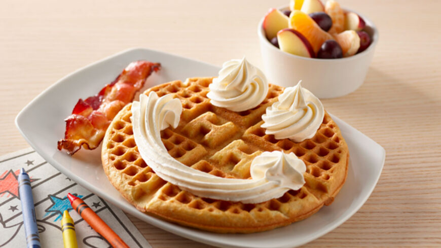 Golden Belgian waffle topped with whipped cream, served with bacon and a bowl of fresh fruit.