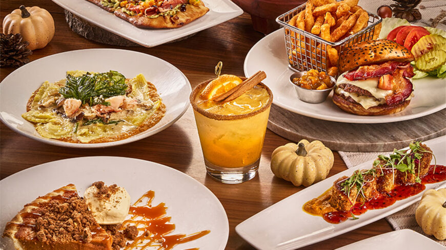 Restaurant spread featuring a bacon cheeseburger with fries, flatbread dishes, a cocktail, and a dessert with ice cream on a wooden table.