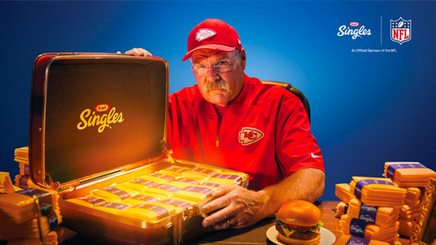 Kansas City Chiefs coach Andy Reid in a red Chiefs cap and jacket holds an open Kraft Singles briefcase filled with individually wrapped cheese slices, with a cheeseburger and stacks of cheese packages on the table.