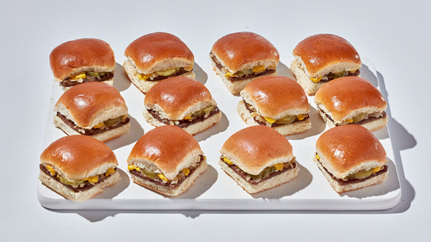 Twelve small cheeseburgers with soft toasted buns, beef patties, cheese, pickles, and onions arranged on a white serving tray.