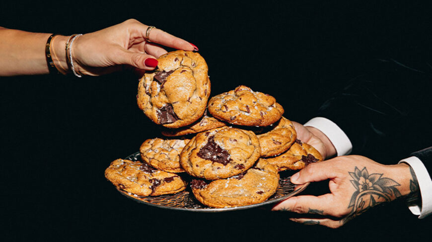 A person holds a chocolate chip cookie over a platter piled with large chocolate chip cookies, with another person holding the platter against a black background.