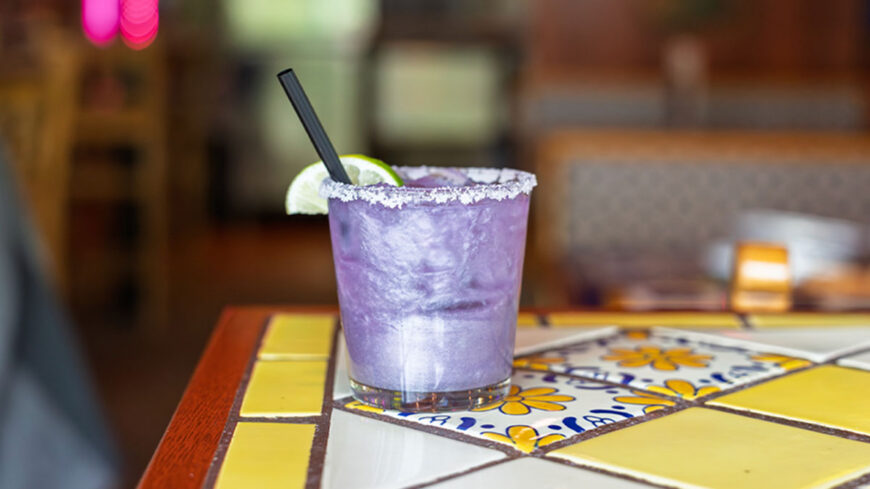 Purple frozen margarita in a salt-rimmed glass with a lime wedge and black straw, set on a colorful tiled restaurant table.