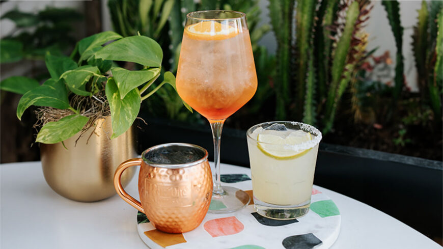 Three cocktails on a patio table—a peach-orange spritz, a pale lime drink in a salted glass, and a copper mug—surrounded by lush green plants.
