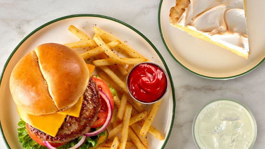 Cheeseburger with lettuce, tomato, onion, and melted cheese served with French fries and ketchup on a plate, with slices of pie and a pale drink nearby.
