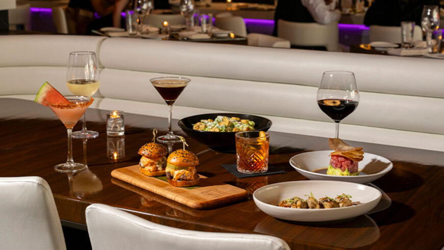 Upscale restaurant dining table featuring cocktails, wine, sliders, appetizers, and plated dishes in a modern, softly lit dining room.