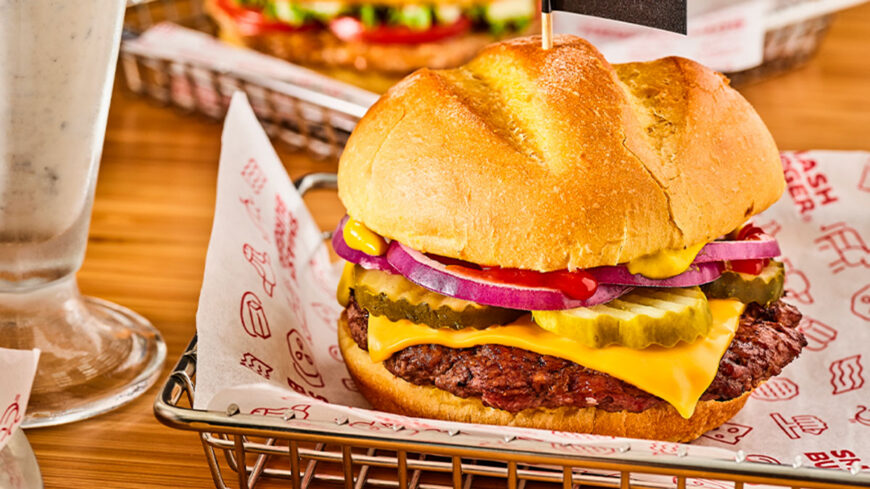 Close-up of a cheeseburger with a toasted bun, beef patty, melted American cheese, pickles, red onion, ketchup, and mustard, served in a basket at a restaurant.