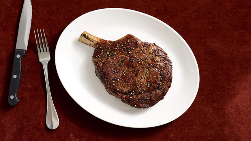Grilled bone-in ribeye steak seasoned with herbs and pepper, served on a white plate with a knife and fork.
