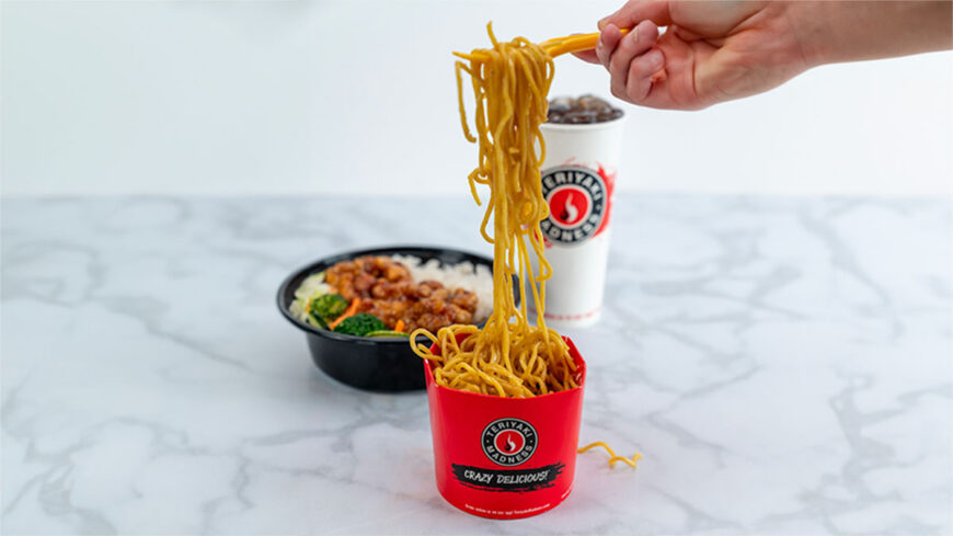 Hand lifting noodles with chopsticks from a red Teriyaki Madness cup, with an orange chicken bowl and branded drink cup in the background.