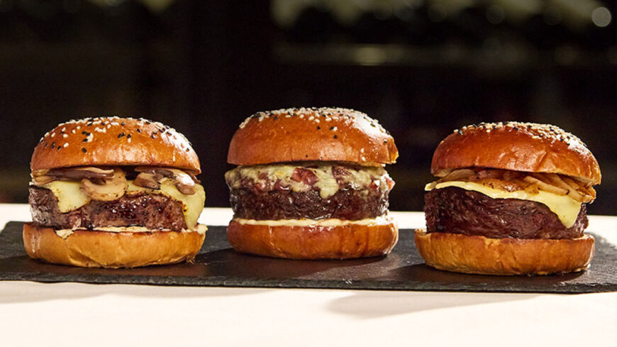 Three gourmet cheeseburgers with toasted sesame-seed buns, thick beef patties, melted cheese, and assorted toppings arranged on a black serving board.