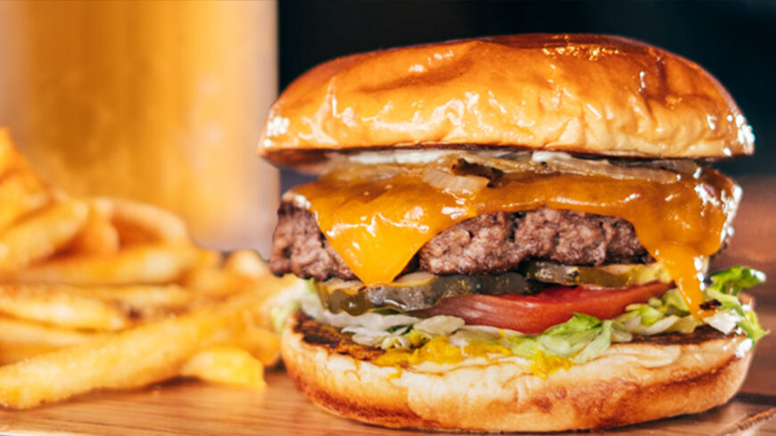 Cheeseburger with melted cheddar cheese, pickles, tomato, lettuce, and onions on a toasted bun, served with fries.