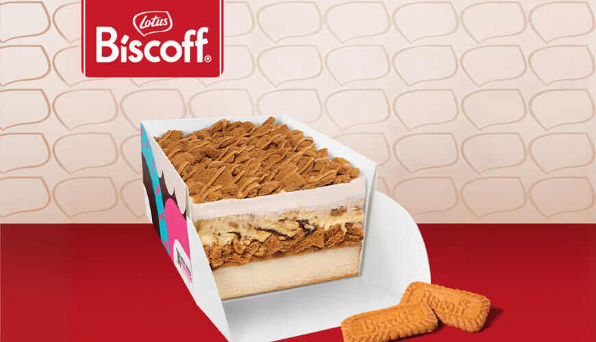 A cross-section view of a Baskin-Robbins Biscoff ice cream cake inside a branded box, showing layers of vanilla cake, ice cream, and a heavy top layer of crushed Biscoff cookies and caramel drizzle.
