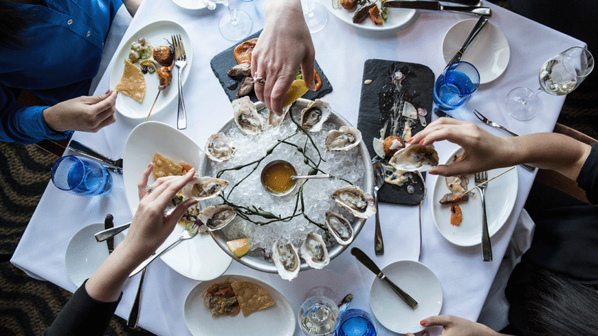 Diners at a restaurant table sharing a platter of oysters on ice, with plates of seafood, drinks, and lemon wedges surrounding it.