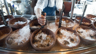 A seafood counter displaying oysters in wooden bowls covered with crushed ice, with a server holding a label identifying the oysters.