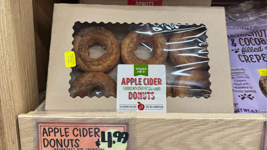 A box of Trader Joe’s apple cider donuts, with several sugar-coated ring-shaped donuts visible through a clear window. A sign below lists the price as $4.99.