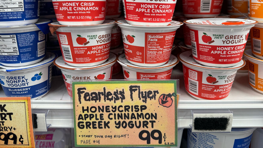Trader Joe’s Honeycrisp Apple Cinnamon Greek Yogurt in red-and-white cups, stacked on a refrigerated shelf. A handwritten sign advertises the 5.3-ounce yogurt for 99 cents.