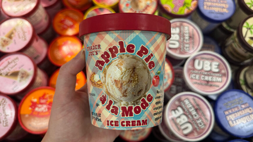 A hand holds a tub of Trader Joe’s Apple Pie La Mode ice cream in front of a freezer filled with assorted ice cream containers.