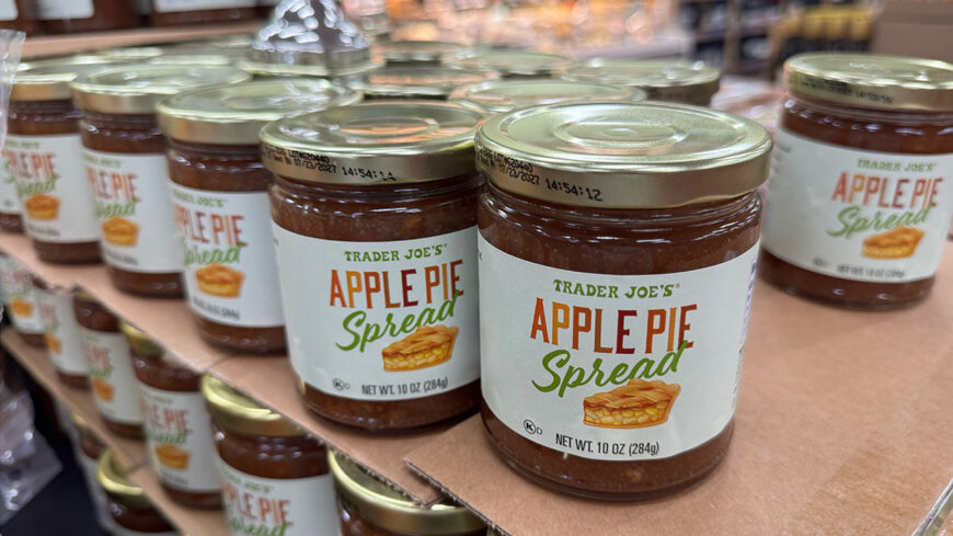 Jars of Trader Joe’s Apple Pie Spread are displayed on wooden shelves, with several jars arranged in rows.