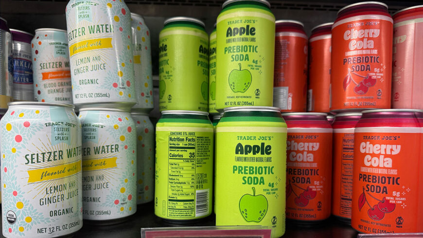 Trader Joe’s Apple Prebiotic Soda and Cherry Cola Prebiotic Soda cans are displayed on a refrigerated shelf alongside lemon-and-ginger seltzer water.