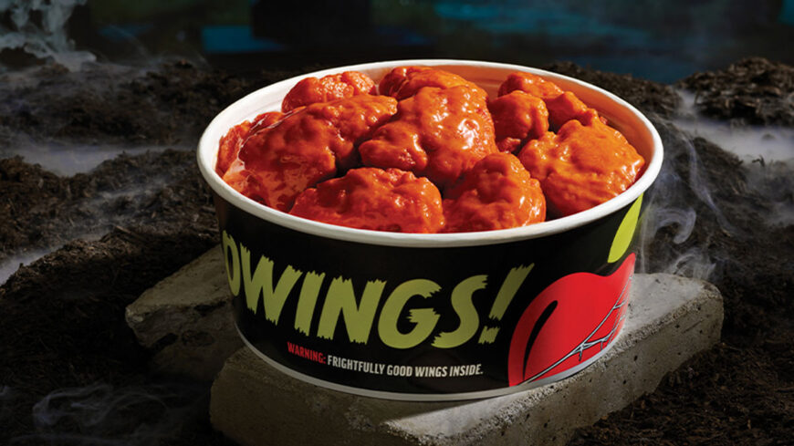 A black Applebee’s Hallowings bucket filled with chicken wings coated in bright red-orange sauce, displayed on a rocky surface surrounded by eerie smoke.