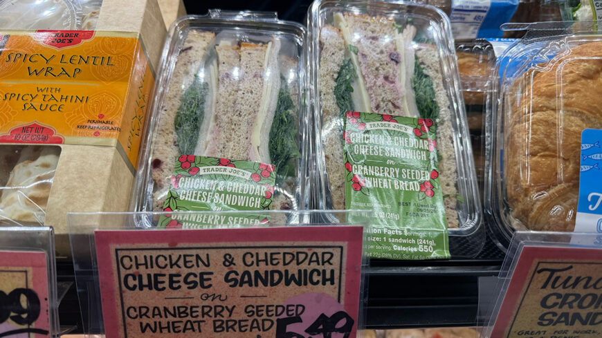 Trader Joe’s chicken and cheddar cheese sandwich on cranberry seeded wheat bread displayed in a refrigerated case, with a spicy lentil wrap visible to the left.