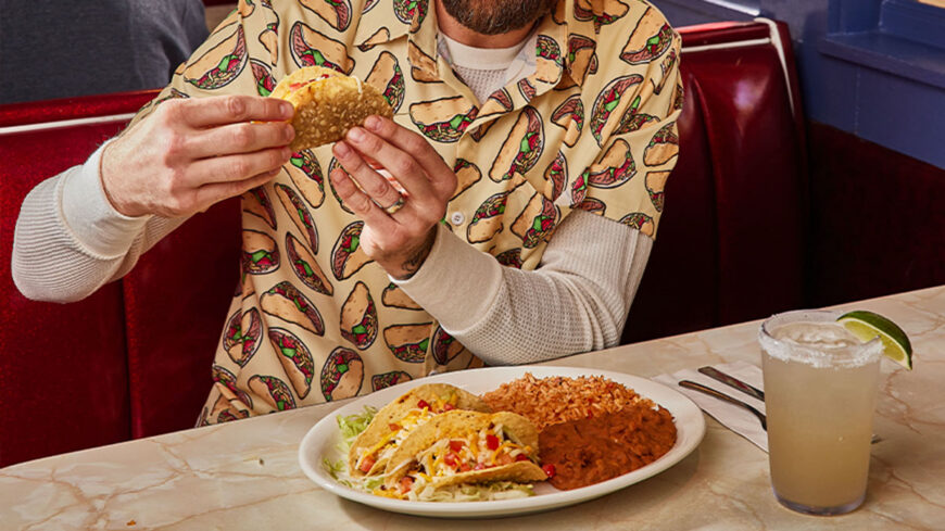 Person holding a crispy taco while seated at a restaurant table with a plate of tacos, rice, beans, and a lime drink.
