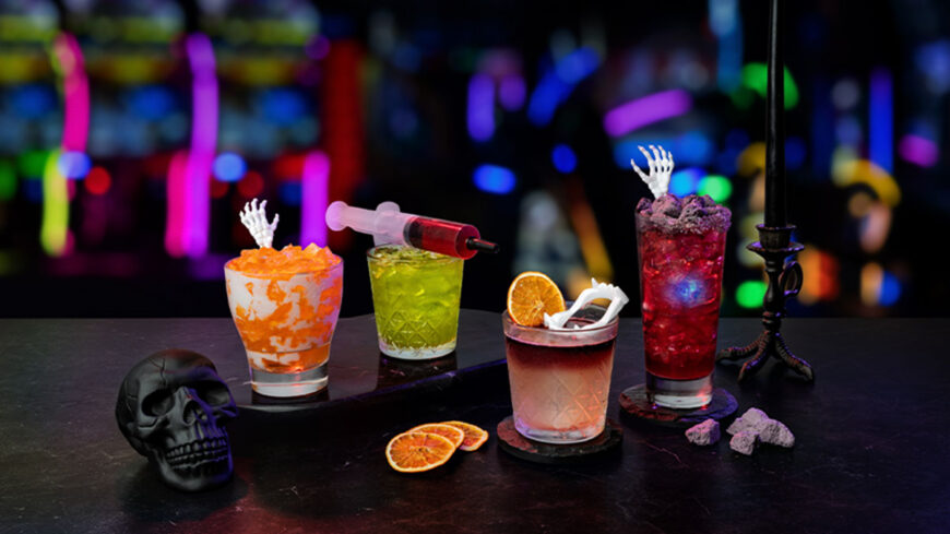 Four colorful Halloween-themed cocktails on a bar, garnished with skeleton hands, crushed candy, and citrus, with a black skull decoration and neon lights in the background.
