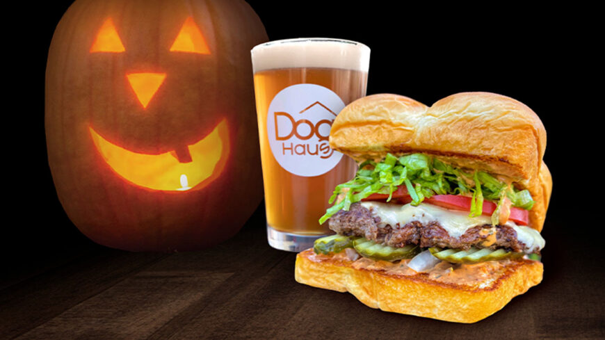A juicy cheeseburger topped with lettuce, tomato, pickles, cheese, and sauce beside a glass of beer, with a glowing carved jack-o’-lantern against a dark background.