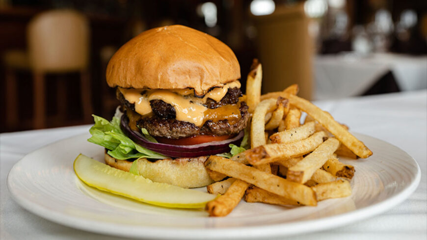 A gourmet double cheeseburger with melted cheese, lettuce, tomato, and red onion, served with golden French fries and a pickle slice on a white plate.