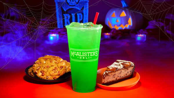 A bright green iced drink in a McAlister’s Deli cup is displayed between a plate of cookies and a slice of chocolate cheesecake on an orange-red surface, with glowing jack-o’-lanterns, blue-purple lighting, and spiderweb decorations in the background.