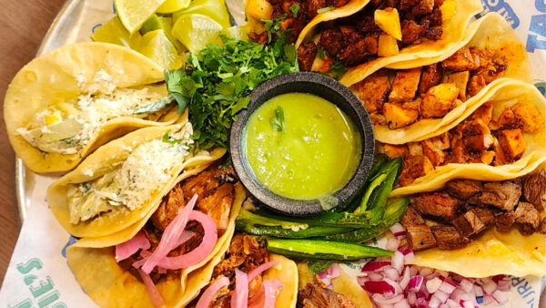 Assorted tacos filled with seasoned chicken, steak, and other meats, served with cilantro, lime wedges, pickled onions, peppers, and green salsa.