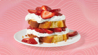 Strawberry shortcake made with layers of golden pound cake, whipped cream, and sliced fresh strawberries, served on a white plate against a pink background with wavy white lines.