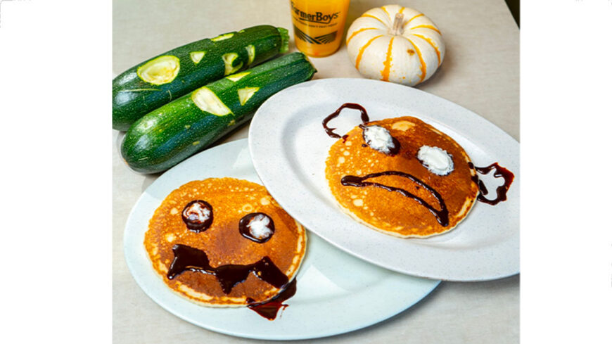 Two golden-brown pancakes decorated with chocolate sauce and whipped cream to look like spooky faces sit on white plates. Green zucchinis and a small white pumpkin are arranged in the background.