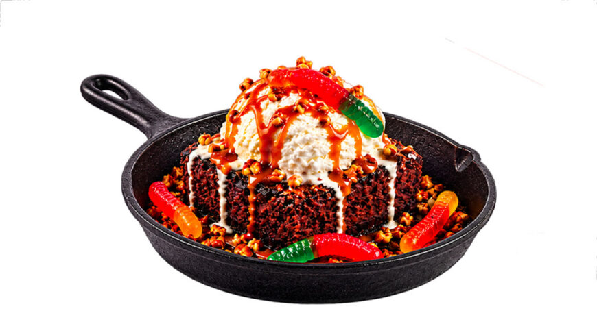 A chocolate brownie sundae topped with vanilla ice cream, caramel and chocolate sauces, candy pieces, and colorful gummy worms is served in a black cast-iron skillet.